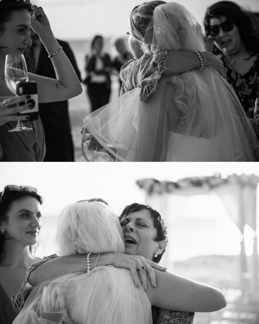 In black and white, guests pull the bride into long hugs after the beach wedding ceremony, a friend beside them holding a wine glass and a phone.
