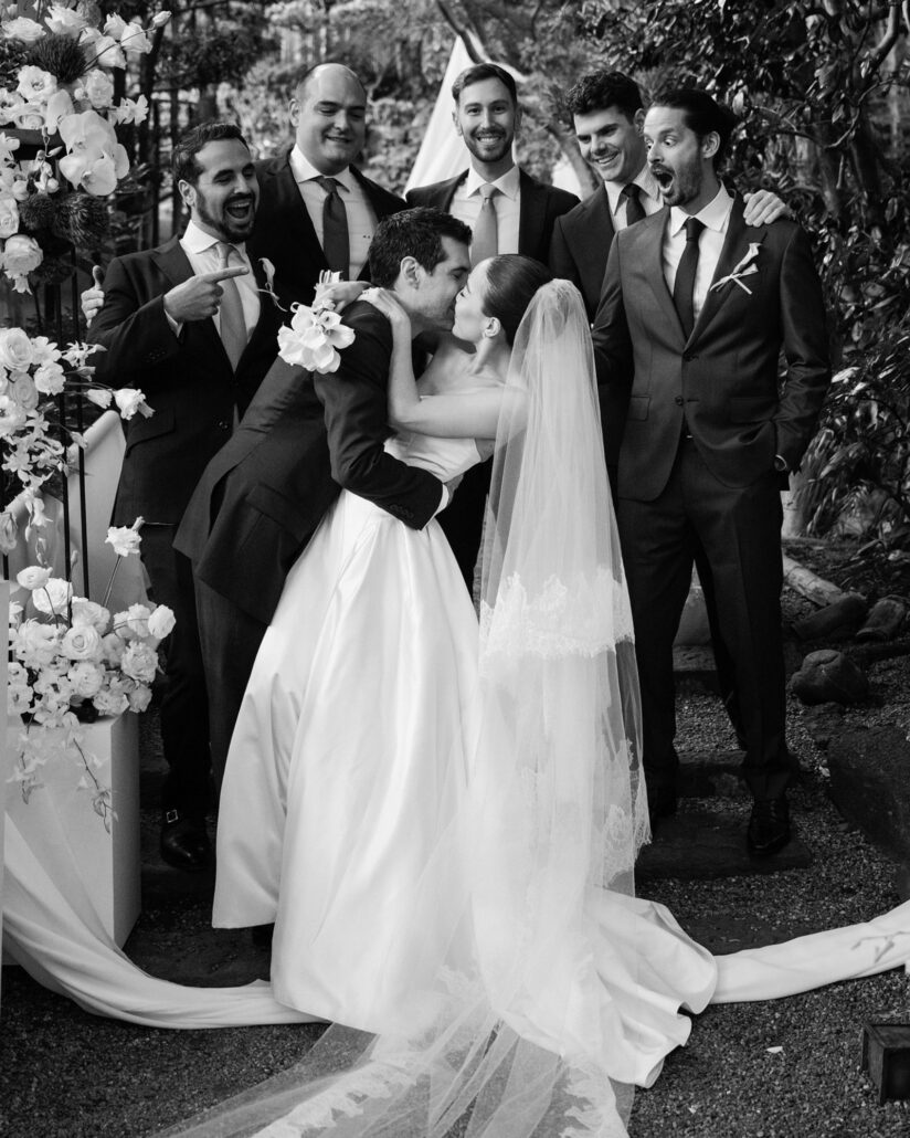 In black and white, the groom leans in to kiss the bride as his groomsmen cheer and point behind them after their Jinya Ryokan garden ceremony.