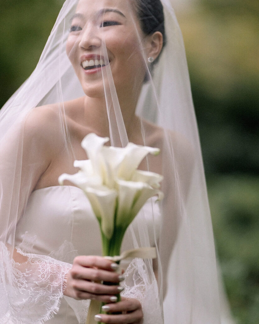 The bride laughs under her veil, holding white calla lilies in the garden at Jinya Ryokan on the day of a Greek and Japanese wedding.