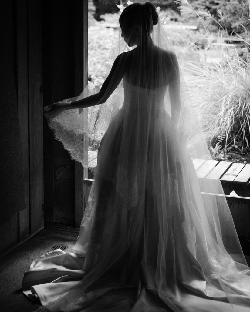 In black and white, the bride stands backlit in a doorway, lifting the lace edge of her veil toward the garden at Jinya Ryokan.