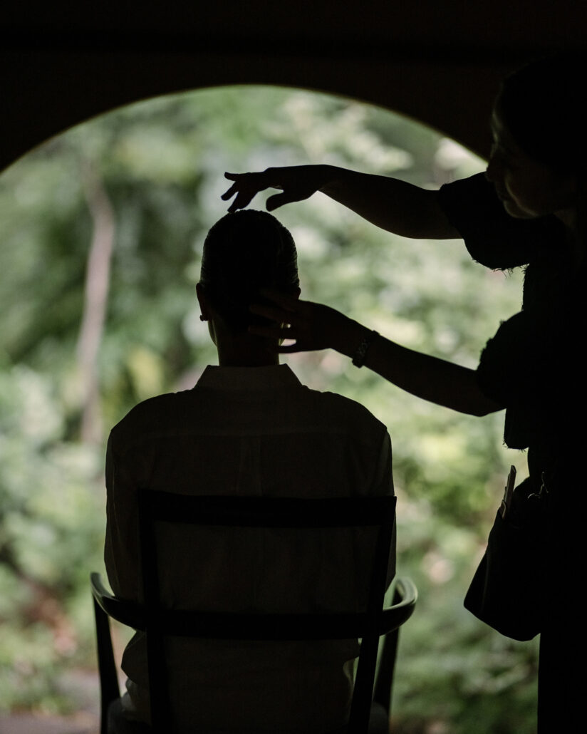 In silhouette, a stylist sets the bride's hair as she sits before a round window full of garden green at Jinya Ryokan.