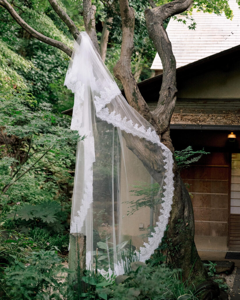 The bride's long veil, edged with lace, hangs from a tree beside the ryokan and lifts in the breeze before a Greek and Japanese wedding at Jinya.
