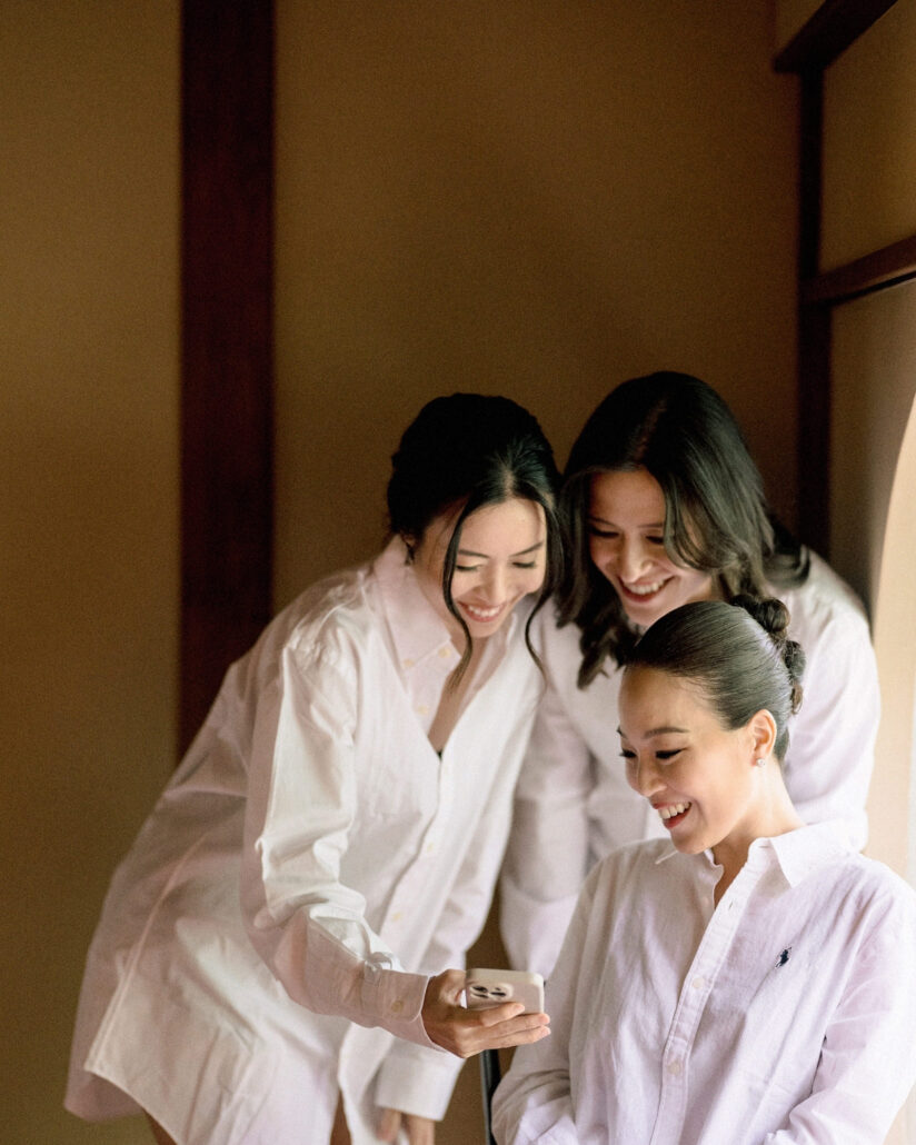 The bride and two friends in white shirts laugh over a phone in a warmly lit room while getting ready for a ryokan wedding in Japan.