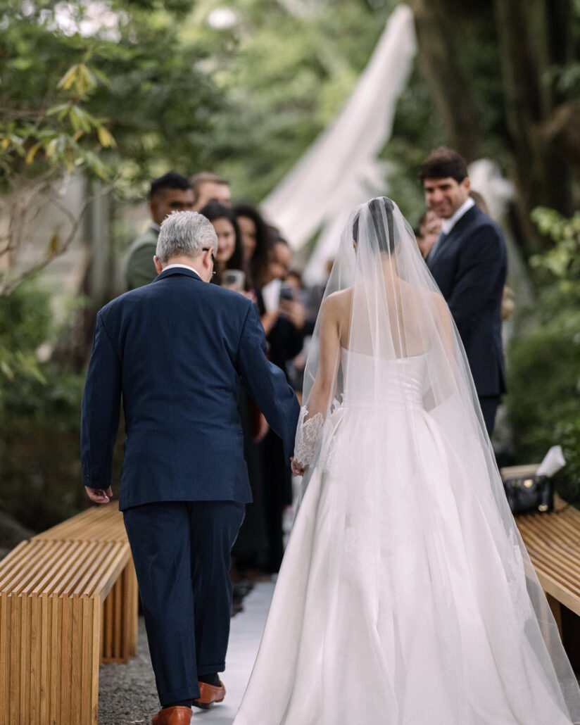The bride walks the garden aisle hand in hand with her father as guests smile and hold up their phones at her Jinya Ryokan wedding.