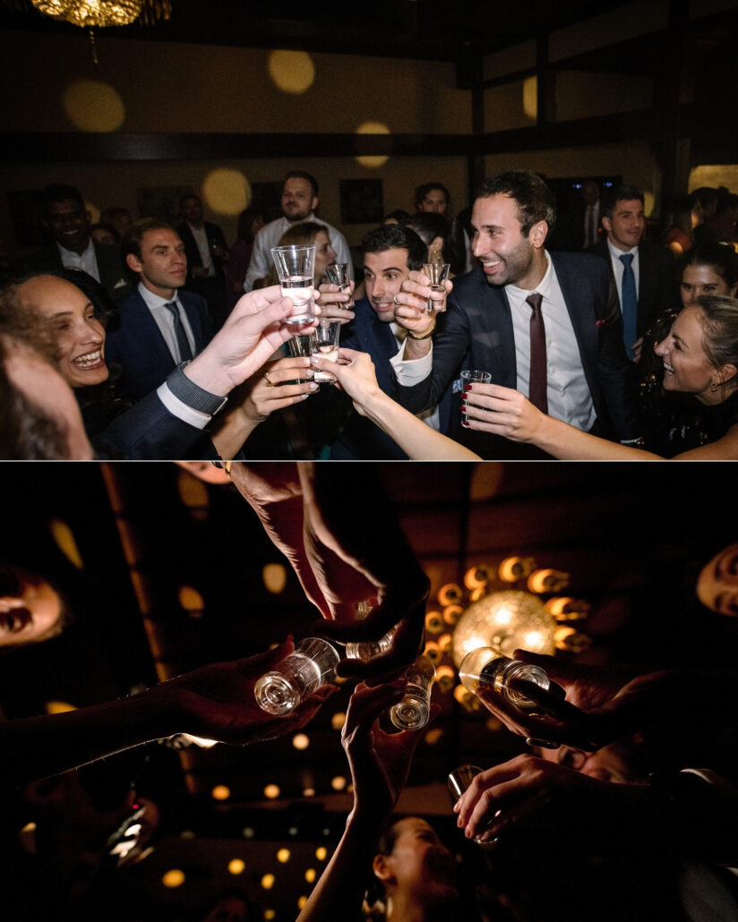Friends raise shot glasses beneath a chandelier, and the glasses meet overhead in the low light of a ryokan wedding reception in Japan.