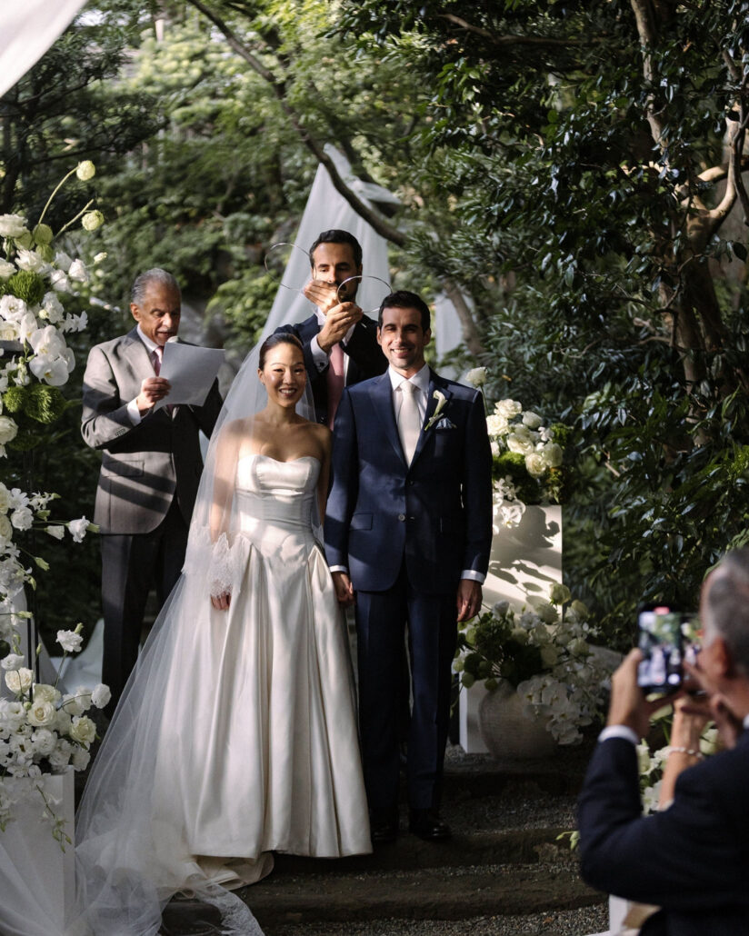 The best man lifts the stefana, two thin Greek wedding crowns, above the smiling couple as the celebrant reads in the garden at Jinya.