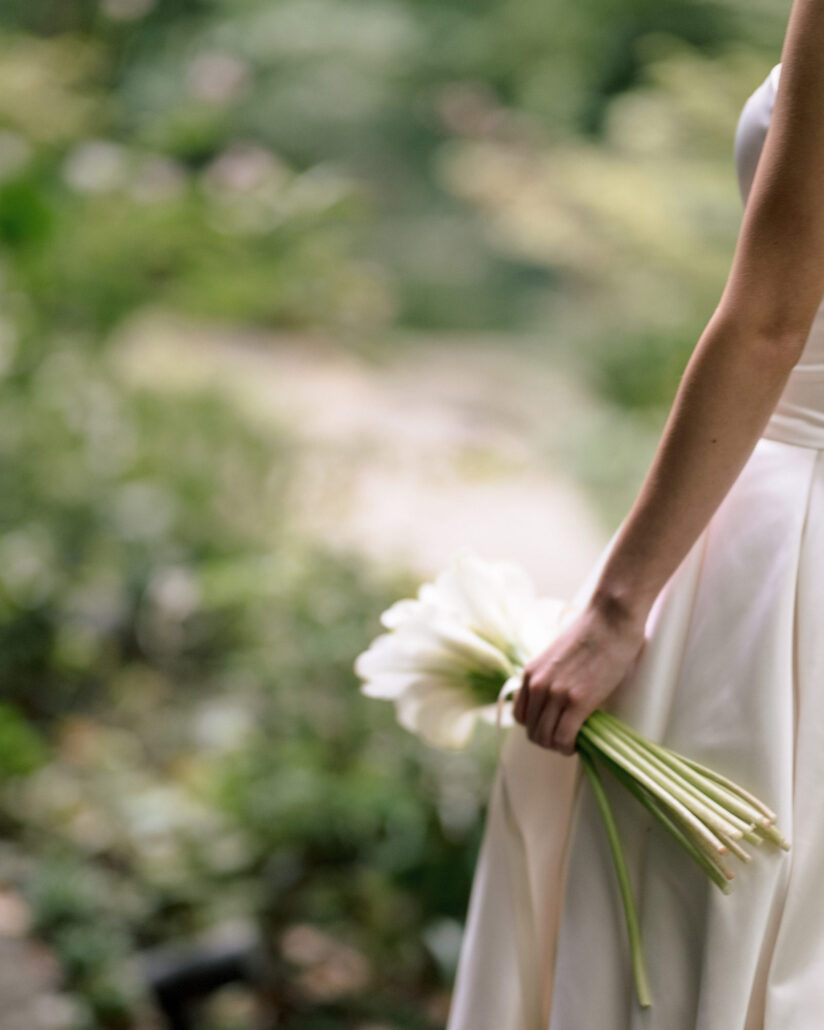 The bride holds a bouquet of white calla lilies loosely at her side against her satin gown in the ryokan garden at Jinya.