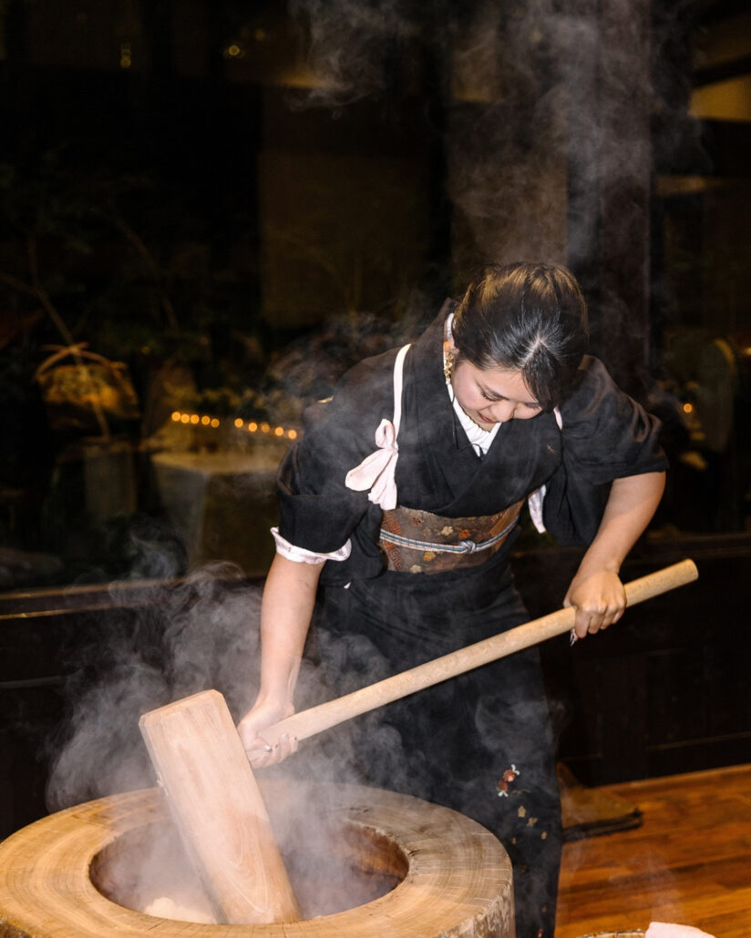 A woman in kimono pounds steaming rice with a wooden mallet for mochi at the reception of a luxury ryokan wedding near Tokyo.