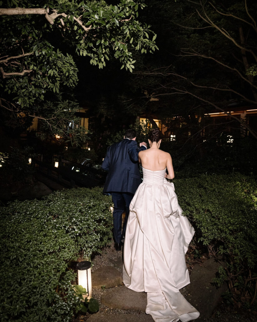 The bride and groom climb a lantern lit stone path into the garden after dark at their ryokan wedding in Japan.