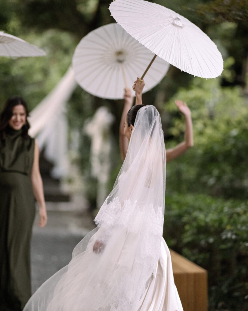 The bride lifts a white paper parasol as a bridesmaid in olive green smiles nearby on the garden path at a Japan destination wedding.