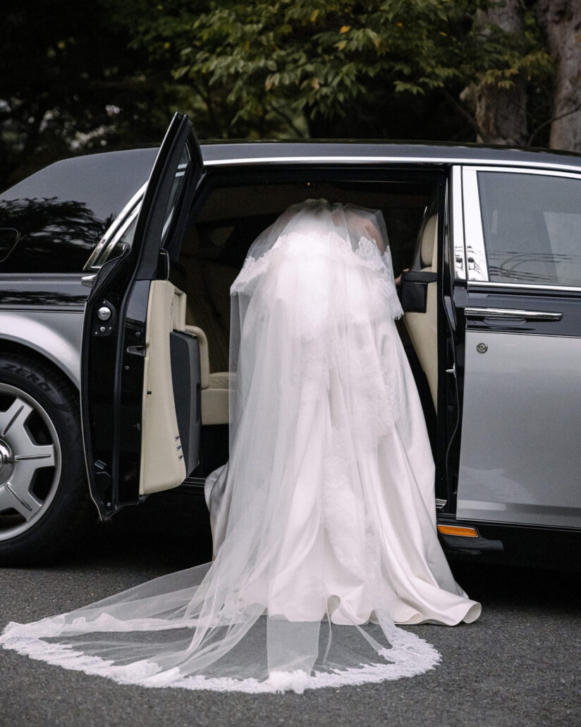 The bride steps into a vintage Rolls Royce as her lace veil spills across the drive at a luxury wedding in Japan.
