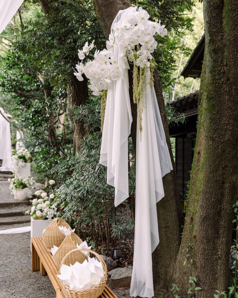 White orchids and draping hang from a tree beside the ceremony aisle, with baskets of folded white paper on a bench at a garden wedding in Japan.