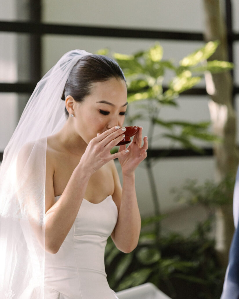 The bride sips from a red lacquer sake cup in a shaded doorway, her veil falling behind her, at a Greek and Japanese wedding in Japan.