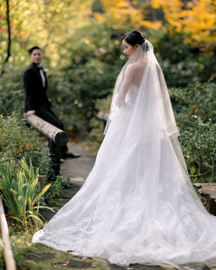 The bride stands in her long veil on a garden path as the groom leans on a log rail behind her, gold leaves above, at a fall wedding in Japan.