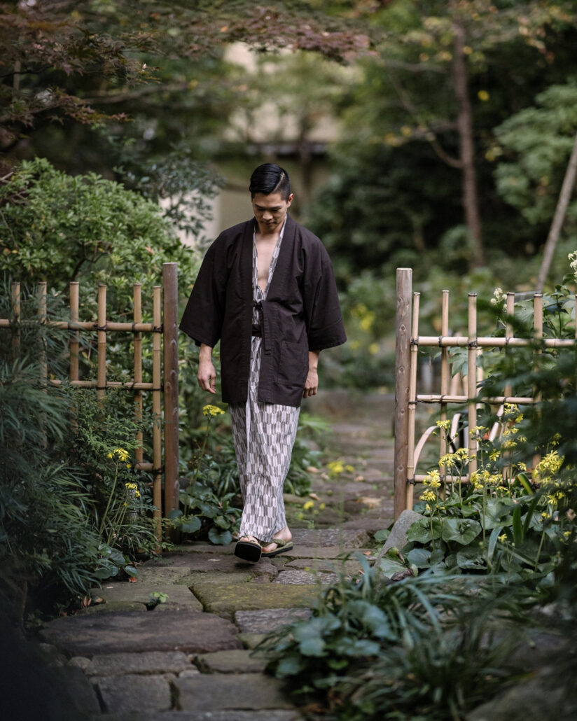 The groom in a yukata walks a stone path through a bamboo gate in the ryokan garden on the morning of his wedding in Japan.