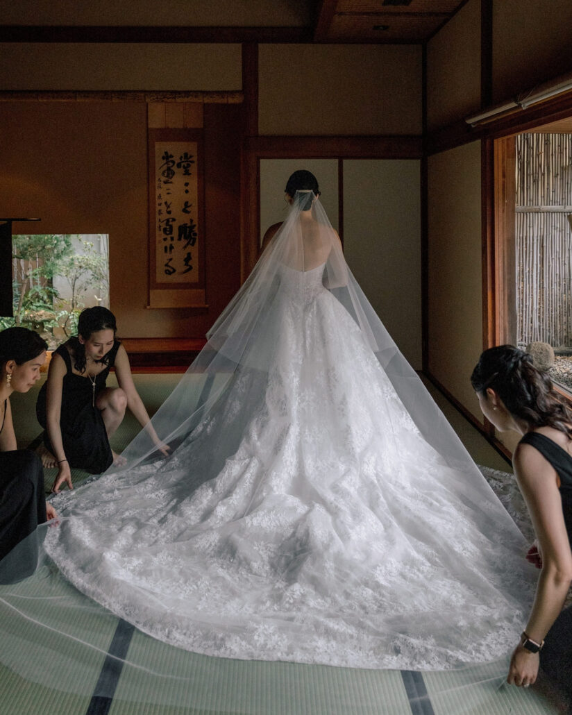 Bridesmaids kneel to spread the bride's long lace train across the tatami as she faces a calligraphy scroll in her suite at a Japanese ryokan.
