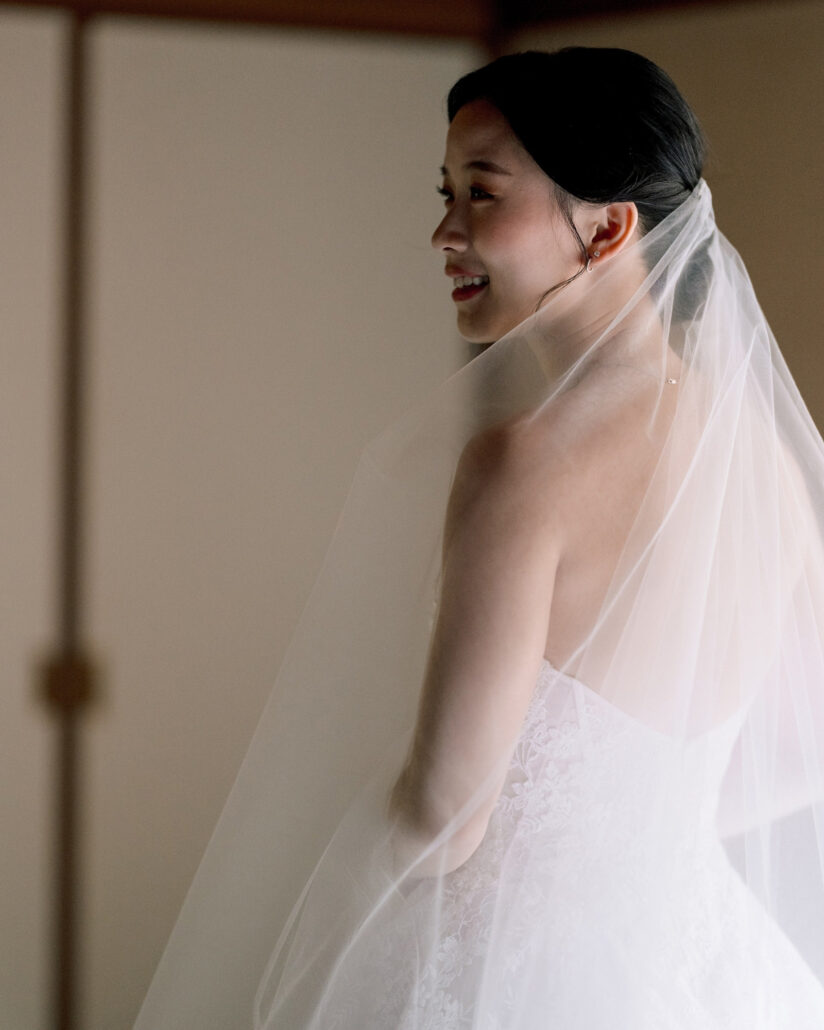 The bride smiles over her shoulder, her sheer veil falling across her back, as she gets ready for her autumn wedding in Japan.