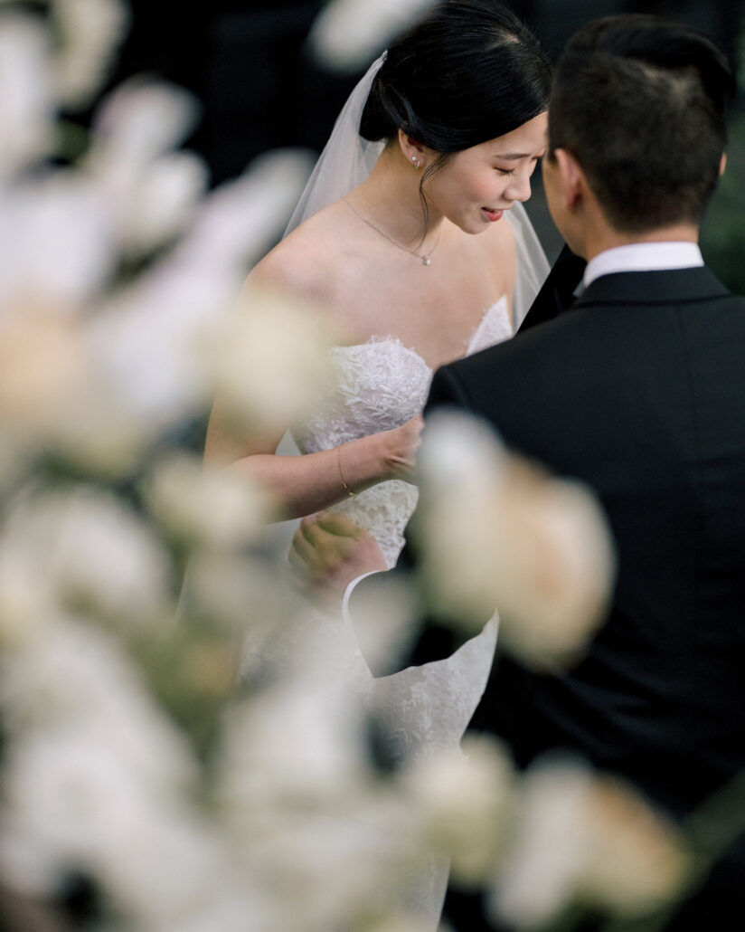 Seen through soft white florals, the bride smiles down as she stands close to the groom during their outdoor wedding ceremony in Japan.