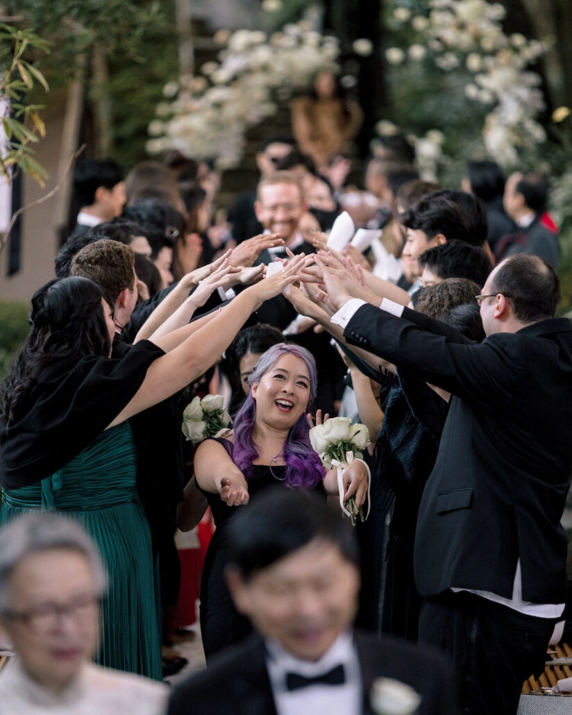 Guests make a tunnel of raised hands over the aisle as a bridesmaid with purple hair and white roses laughs her way through at an outdoor wedding in Japan.