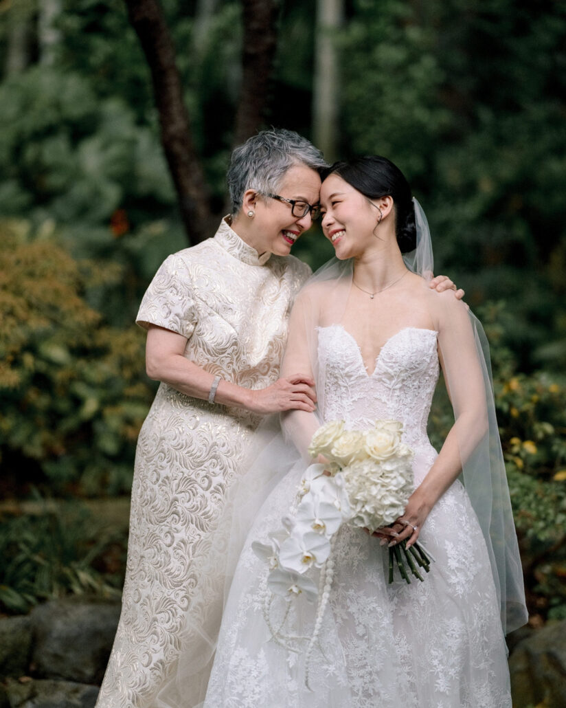 The bride and her mother rest their foreheads together and laugh in the green garden of the ryokan on her wedding day in Japan.
