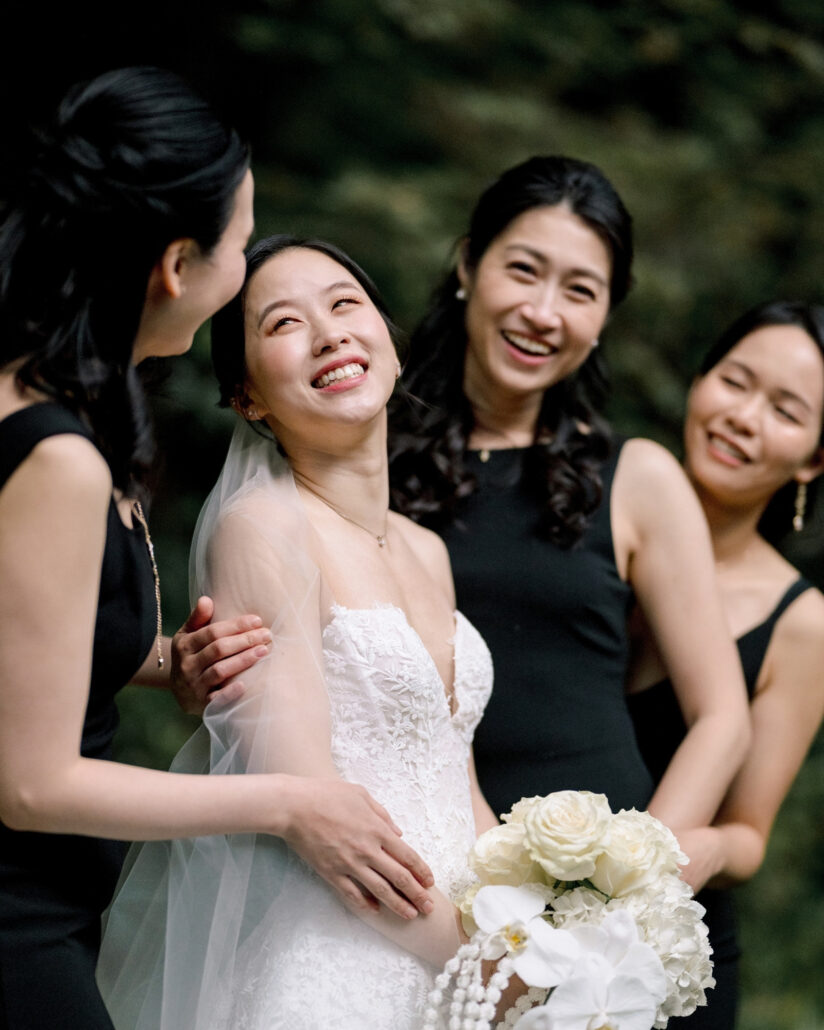 The bride laughs and looks up as her bridesmaids in black gather close in the garden at her destination wedding in Japan.