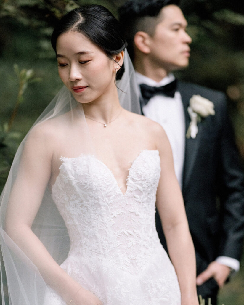 The bride lowers her eyes, her veil falling past her shoulder, as the groom looks away behind her in the green garden of their ryokan wedding in Japan.