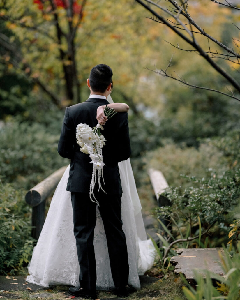 Seen from behind, the groom holds the bride as her arms circle his neck, her cascading orchid bouquet down his back at an autumn garden wedding in Japan.