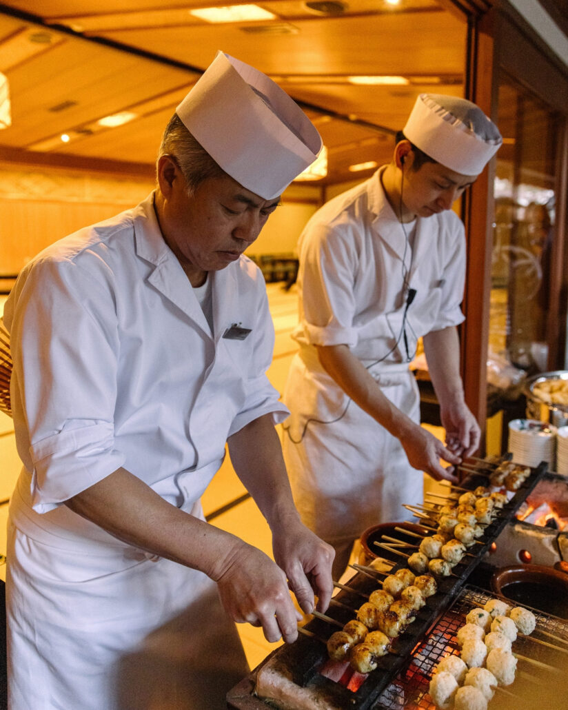 Two chefs in white caps grill yakitori skewers over glowing charcoal at a ryokan buyout wedding at Jinya in Japan.