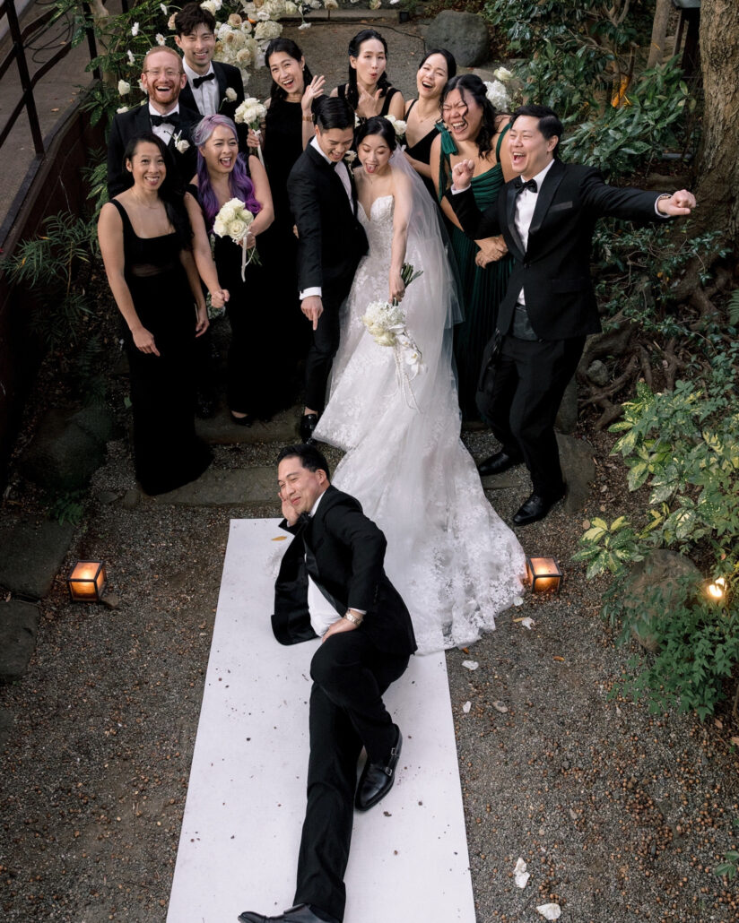 From above, a friend in a tuxedo lies on the white aisle runner as the couple and their wedding party laugh at their Japanese garden wedding.