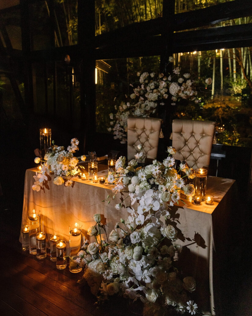 Candles and white florals line the sweetheart table for the wedding reception in Japan, with a lit bamboo grove beyond the windows.