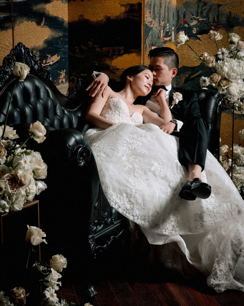 The bride rests against the groom on a black leather sofa among white roses and gold folding screens in the photo booth of a luxury wedding in Japan.