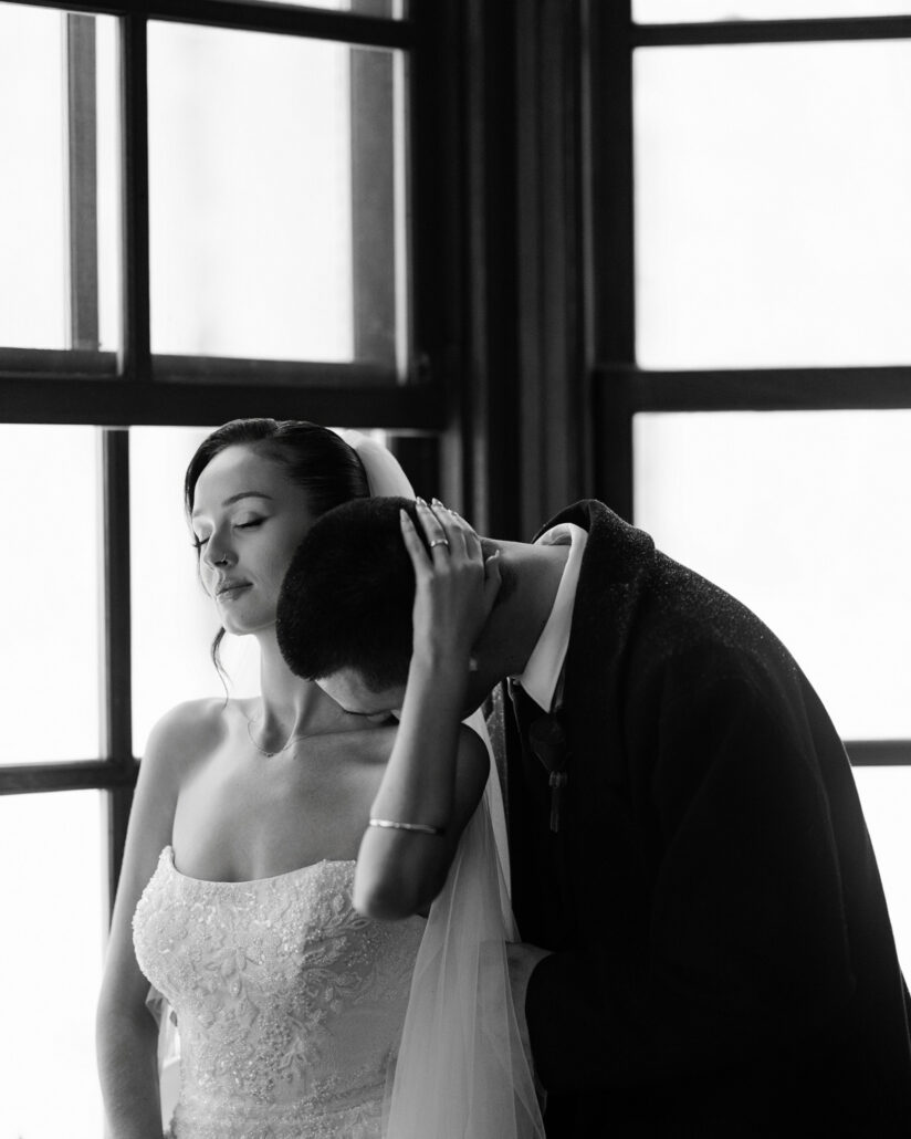 In black and white, the groom rests his head on the bride's shoulder by a tall window, her hand on the back of his head, on their wedding day in Japan.