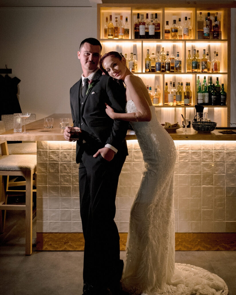 The bride leans into the groom at the backlit whisky bar during cocktail hour, shelves of bottles glowing behind them in Hakuba.