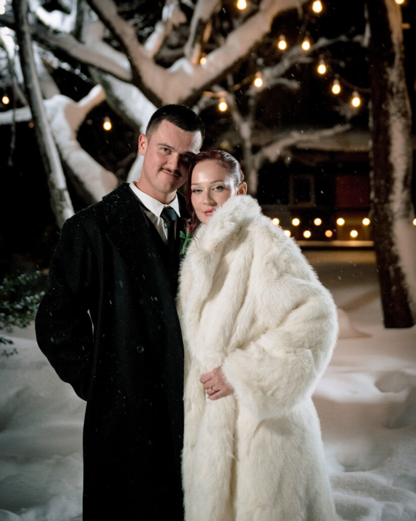 The groom and the bride in a long white fur coat stand close under string lights and snow laden branches at night after their snow wedding in Hakuba.