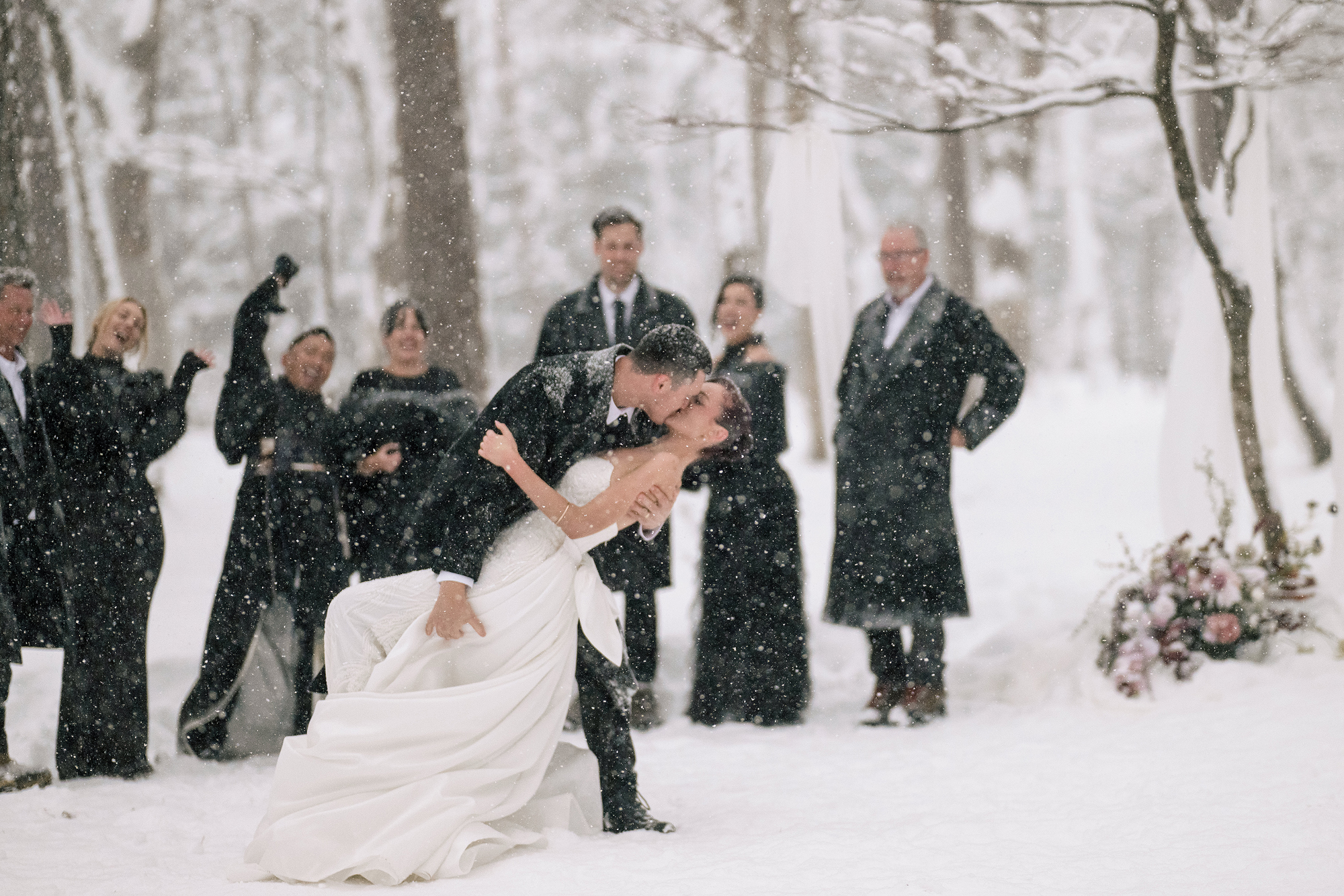 The groom dips the bride for a kiss in heavy falling snow as the wedding party cheers in the forest at their Hakuba snow wedding.