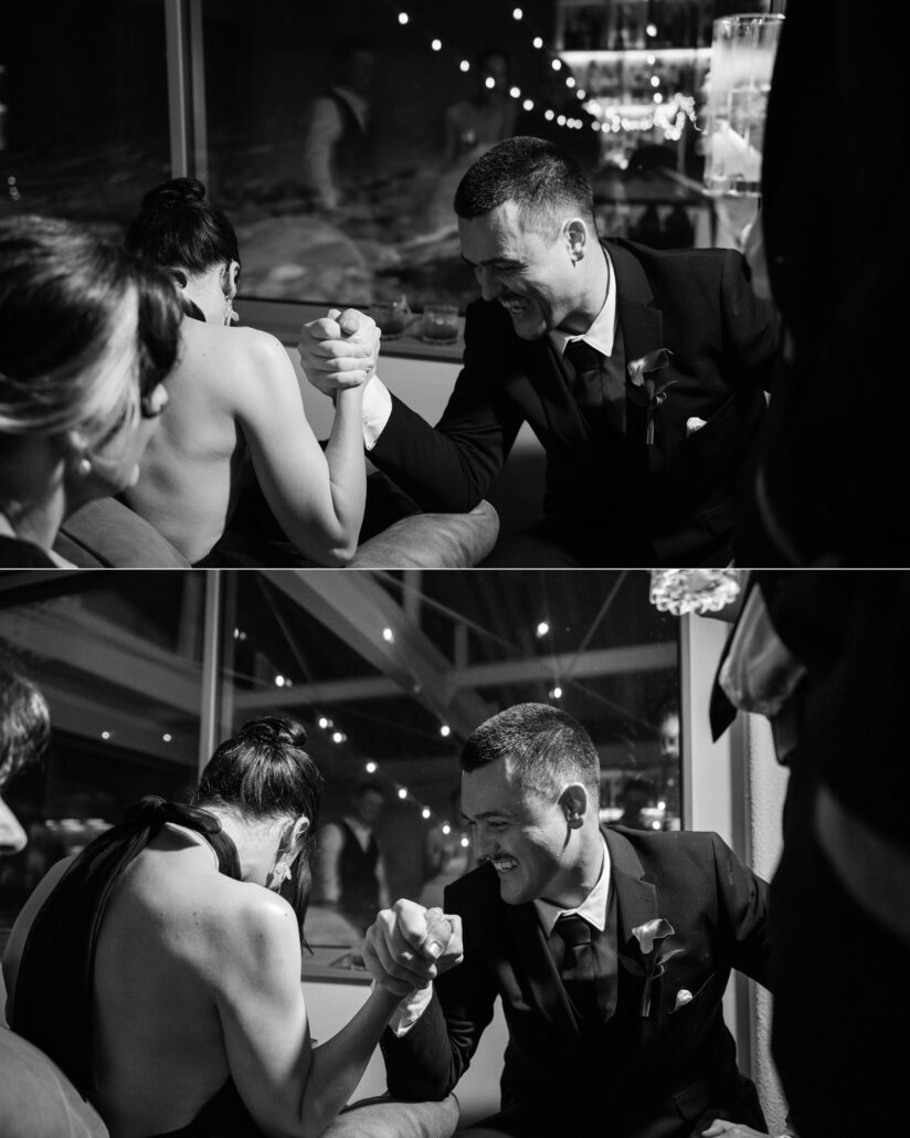 In black and white, a friend in a backless black top arm wrestles a laughing man in a dark suit under string lights at the Hakuba wedding party.