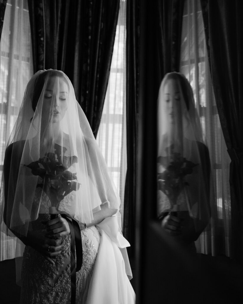 In black and white, the bride stands beneath her veil holding dark calla lilies, her reflection caught in the window glass on her winter wedding day.