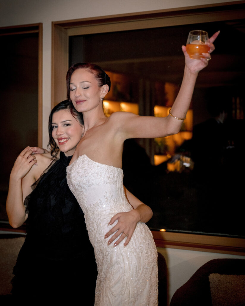 The bride raises her glass high with her eyes closed as a friend in black hugs her close in the warm light of the whisky bar at her Hakuba winter wedding.
