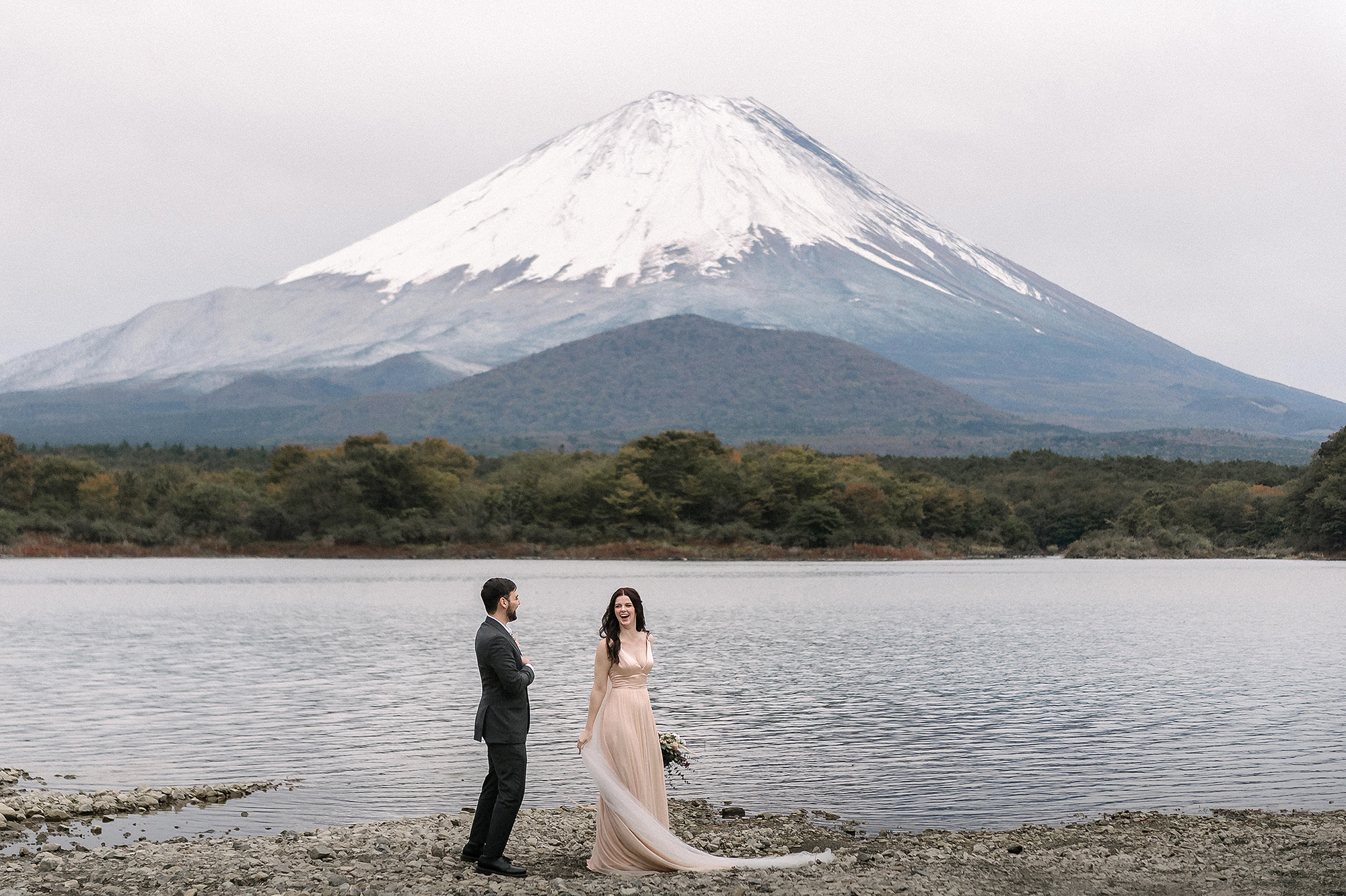 The bride laughs in a blush gown and long sheer train as the groom faces her on a lakeshore below snow capped Mt Fuji, a destination wedding in Japan.