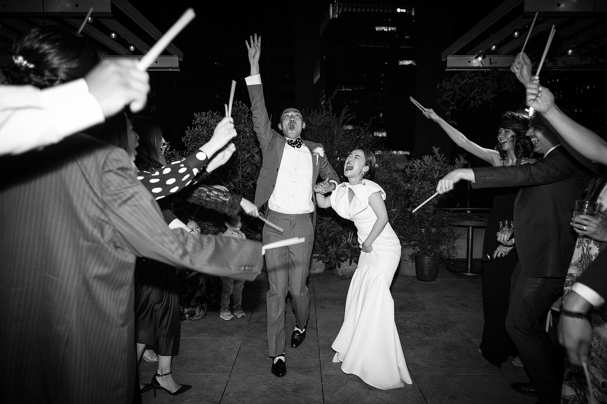 In black and white, the groom throws an arm up and the bride laughs as they run through a tunnel of guests waving white sticks at a Tokyo rooftop wedding.