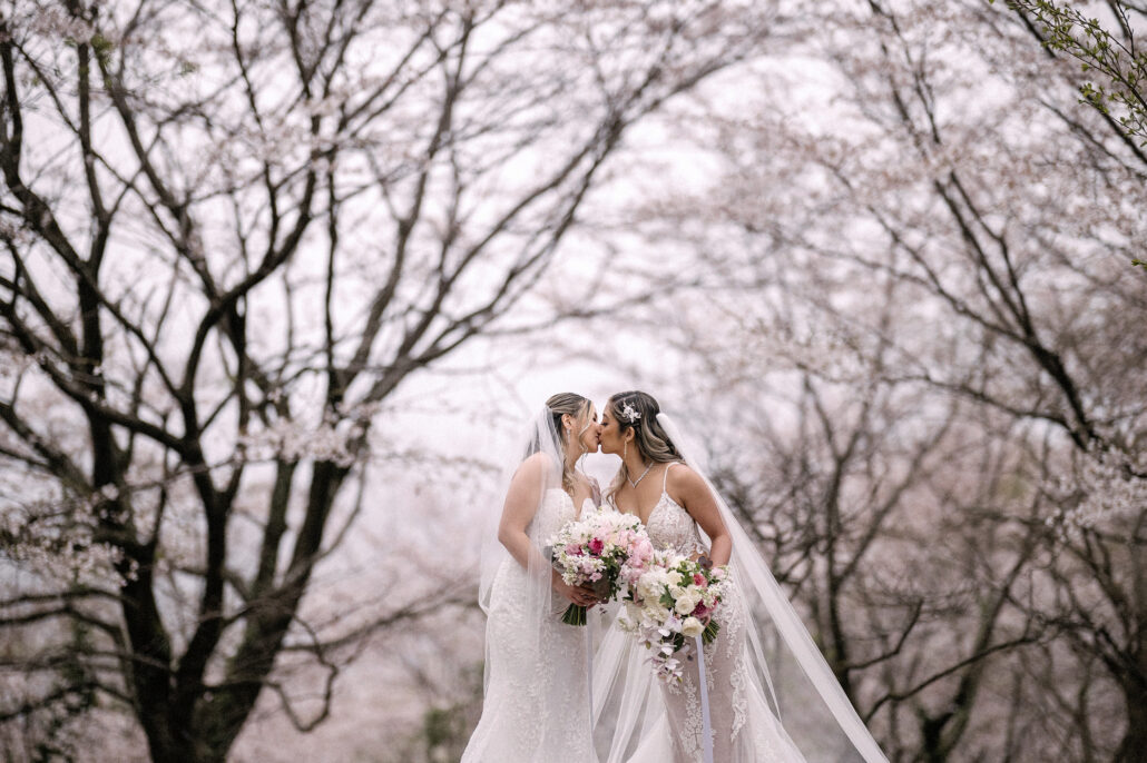 Two brides in lace gowns and long veils kiss beneath pale cherry blossom, holding bouquets of pink and white flowers, an LGBTQ wedding in Japan.