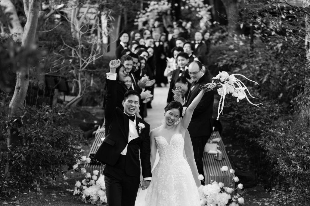 In black and white, the newlyweds walk back down a garden aisle with their arms raised as their guests cheer behind them.