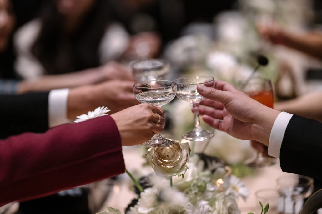 Wedding guests raise coupe glasses in a toast across a reception table of white roses and daisies.