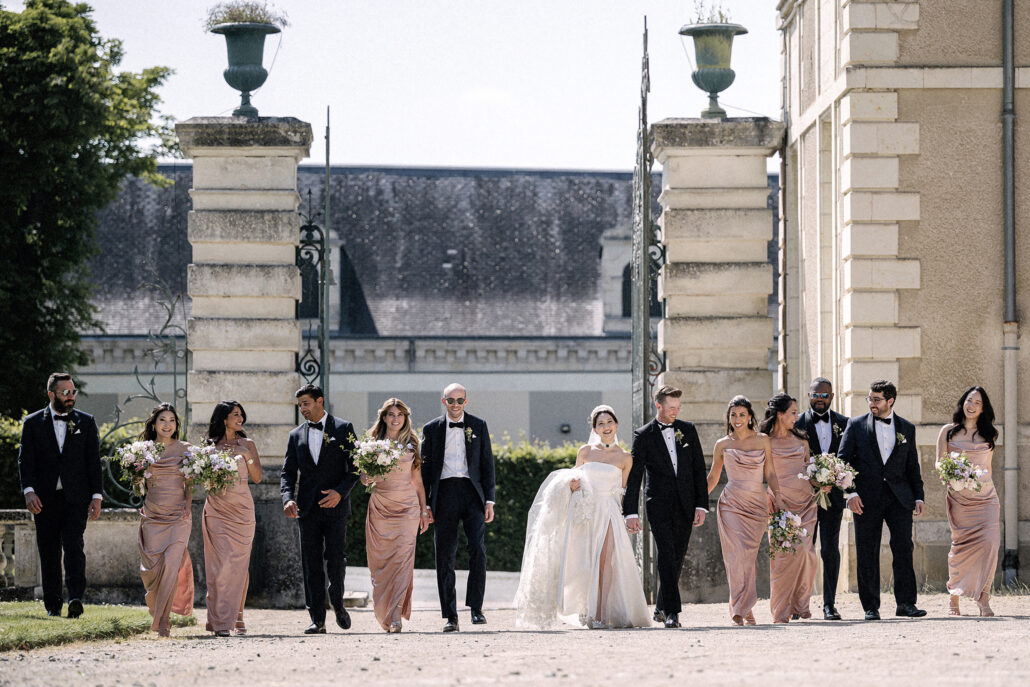 The bride and groom walk through the iron gates of a stone chateau with their bridesmaids in blush satin and groomsmen in black tie.