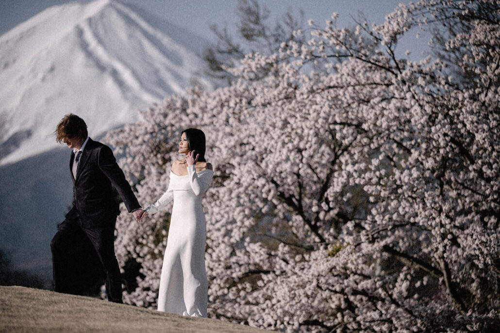 The couple walk hand in hand along a hillside of cherry blossom in full bloom, the snow-covered slopes of Mt Fuji behind them, in mid April.