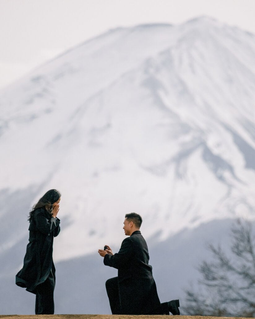 He kneels with the ring as she covers her face in surprise, a snow-capped Mount Fuji filling the sky behind their Kawaguchiko proposal.