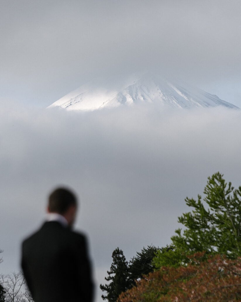 The groom stands with his back to the camera as the snowy summit of Mt Fuji appears above the cloud on a spring elopement morning at Lake Kawaguchi.