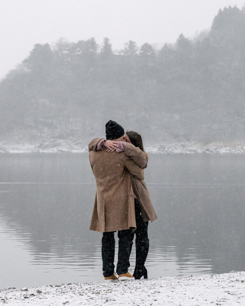 The newly engaged couple hold each other on the snowy shore of Lake Shoji as snow falls over the misty hills, a winter proposal in Japan.