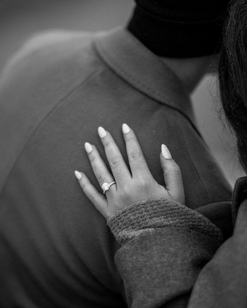 Her new engagement ring up close as her hand rests on his winter coat, in black and white.