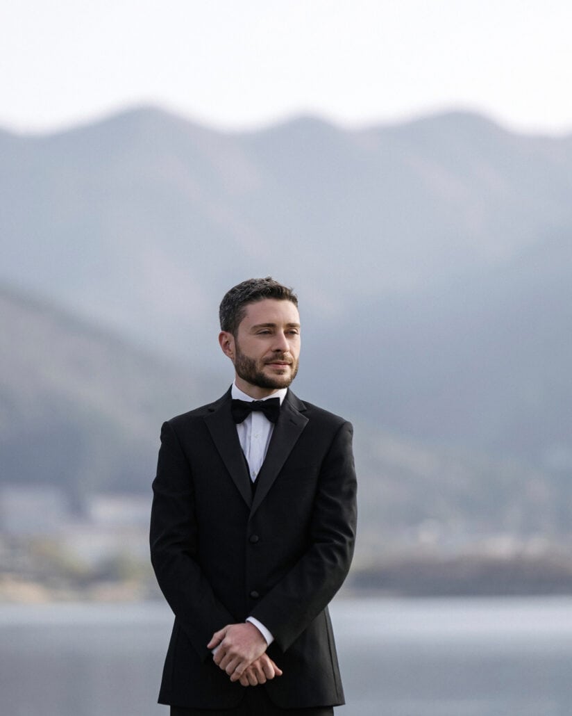 The groom in a black tuxedo and bow tie waits on the shore of Lake Kawaguchi for the first look, hands clasped, the mountains behind him.